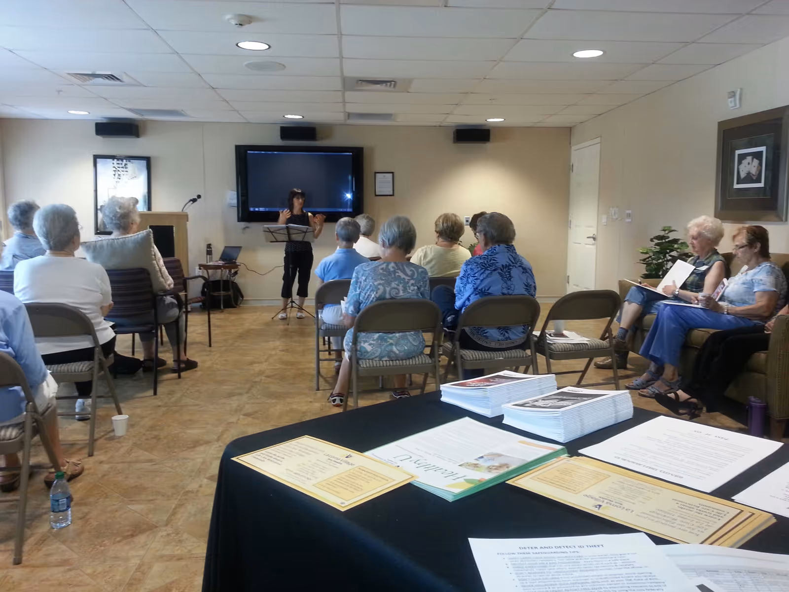 A group of elderly people seated in a room facing a woman playing a violin near a large screen on the wall. Some attendees are seated on chairs, while others are on a couch. A table in the foreground holds various papers and brochures.