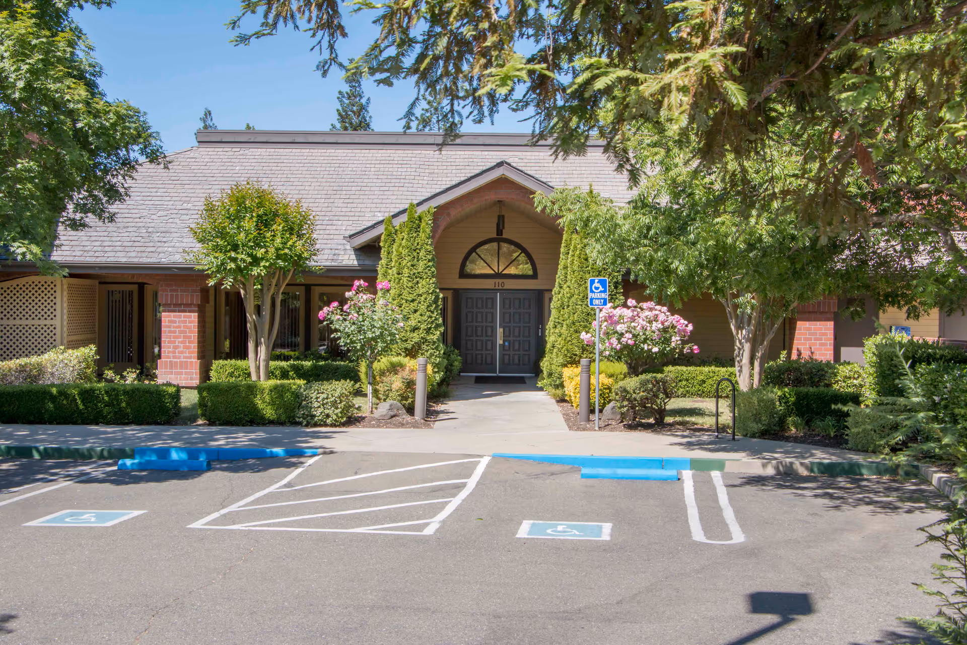 Front exterior view of a single-story building with a peaked roof, surrounded by trees and bushes. There is a paved parking area in front with two handicap parking spaces marked in blue. The entrance has double doors under a small covered porch with two tall shrubs on either side and flowering bushes nearby.