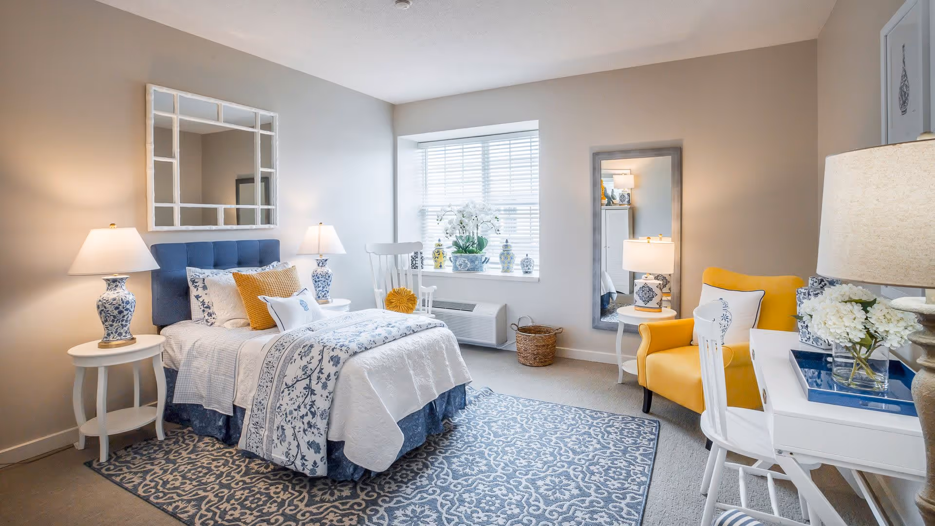 A bright and cozy bedroom with a single bed dressed in white and blue patterned bedding, two bedside tables with matching blue and white lamps, a large window with white blinds, a yellow armchair, a white desk with a chair, a full-length mirror, and decorative vases on the windowsill.