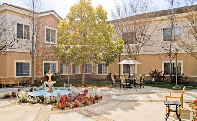 Outdoor courtyard area at a senior living facility with a central water fountain surrounded by colorful plants, several benches, a table with chairs and an umbrella, and a two-story building with windows in the background.