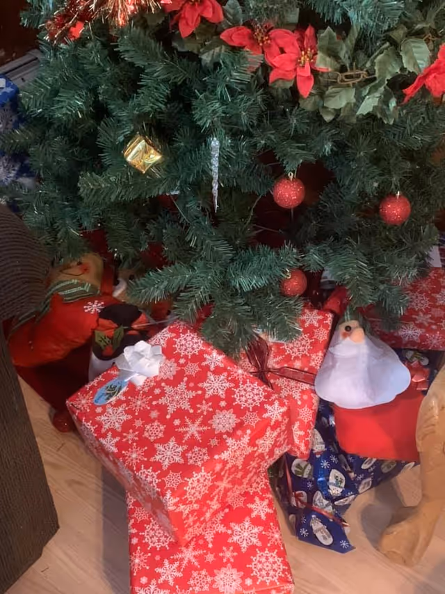 Christmas tree decorated with red poinsettias, red baubles, and a small gold gift ornament. Underneath the tree are several wrapped presents in red and blue wrapping paper with snowflake and snowman patterns. There is also a small Santa Claus decoration and a white bird decoration among the gifts.