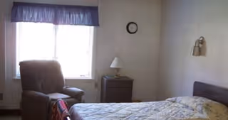 Bedroom with a bed topped by a patterned quilt, a nightstand with a lamp, a recliner by a window, and a clock on the wall.
