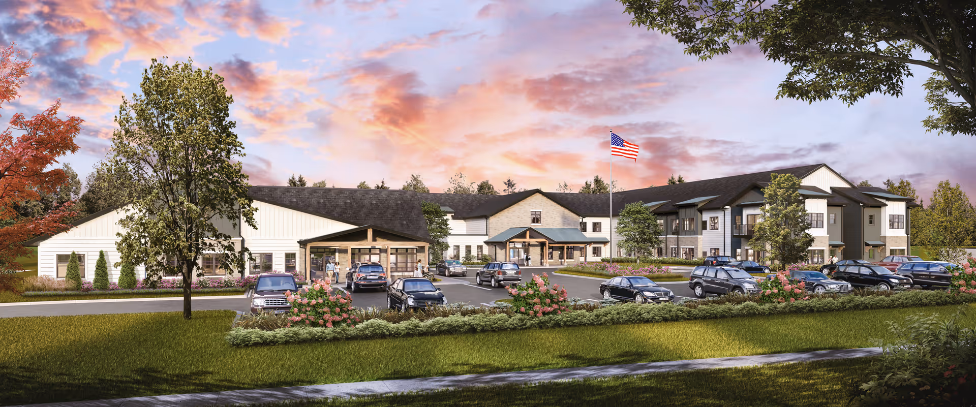Front exterior of a two-story assisted living facility with landscaped grounds, parked cars, and an American flag under a colorful sky.