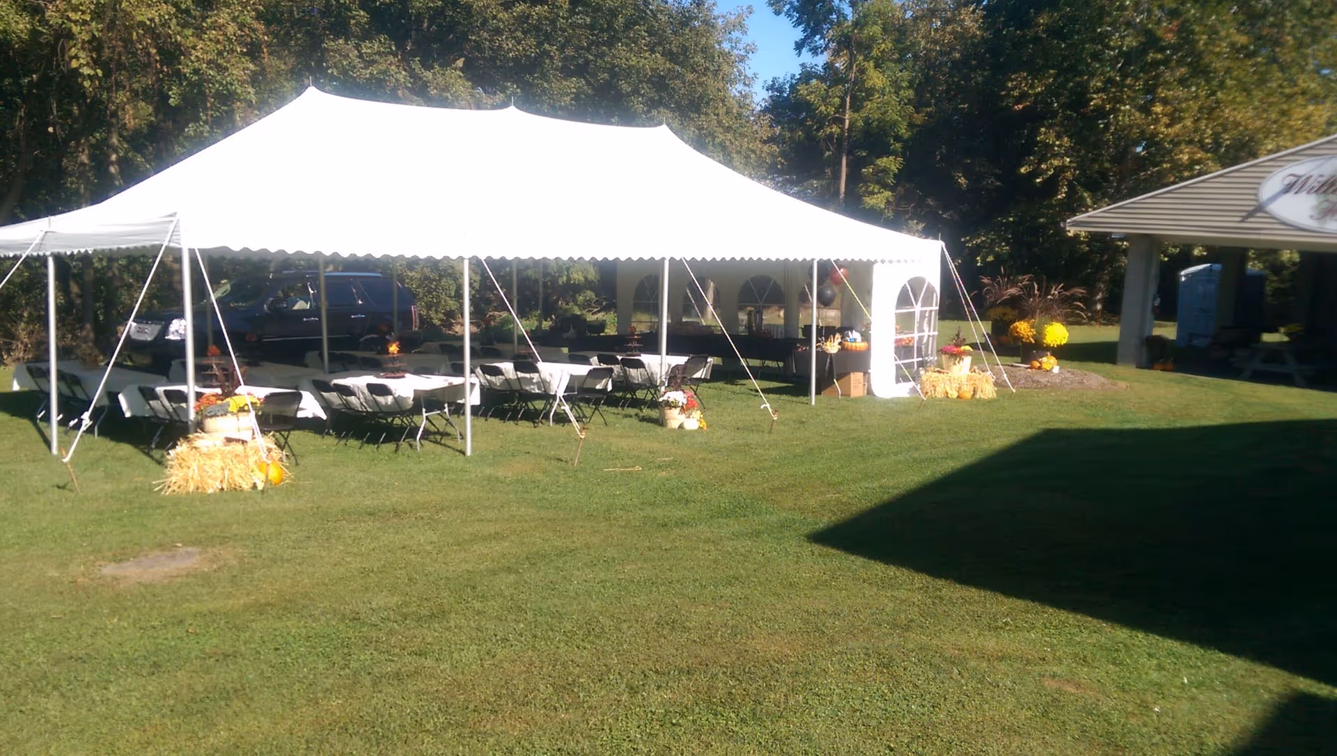 A large white event tent set up on a grassy lawn with rows of tables and chairs underneath. There are hay bales and flower arrangements around the tent, and trees in the background. To the right, there is a covered pavilion with a sign that reads 'Willow View Home'.