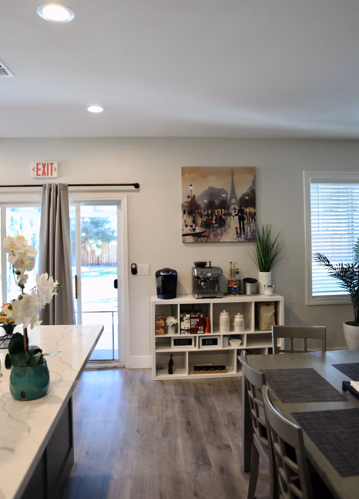 Interior view of a room in Rocklin Care Home showing a coffee station with a coffee maker, espresso machine, and various coffee supplies on a white shelf. There is a dining table with chairs and placemats on the right side. A sliding glass door with curtains leads outside, and a painting of a Paris street scene with the Eiffel Tower hangs on the wall above the coffee station. The floor is wood, and there are potted plants near the window with blinds.