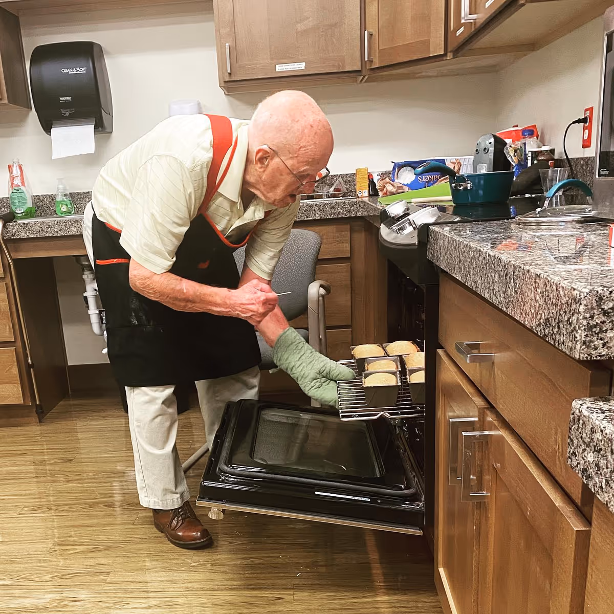 An older man in an apron uses an oven mitt to remove small loaves from an open oven in a communal kitchen.
