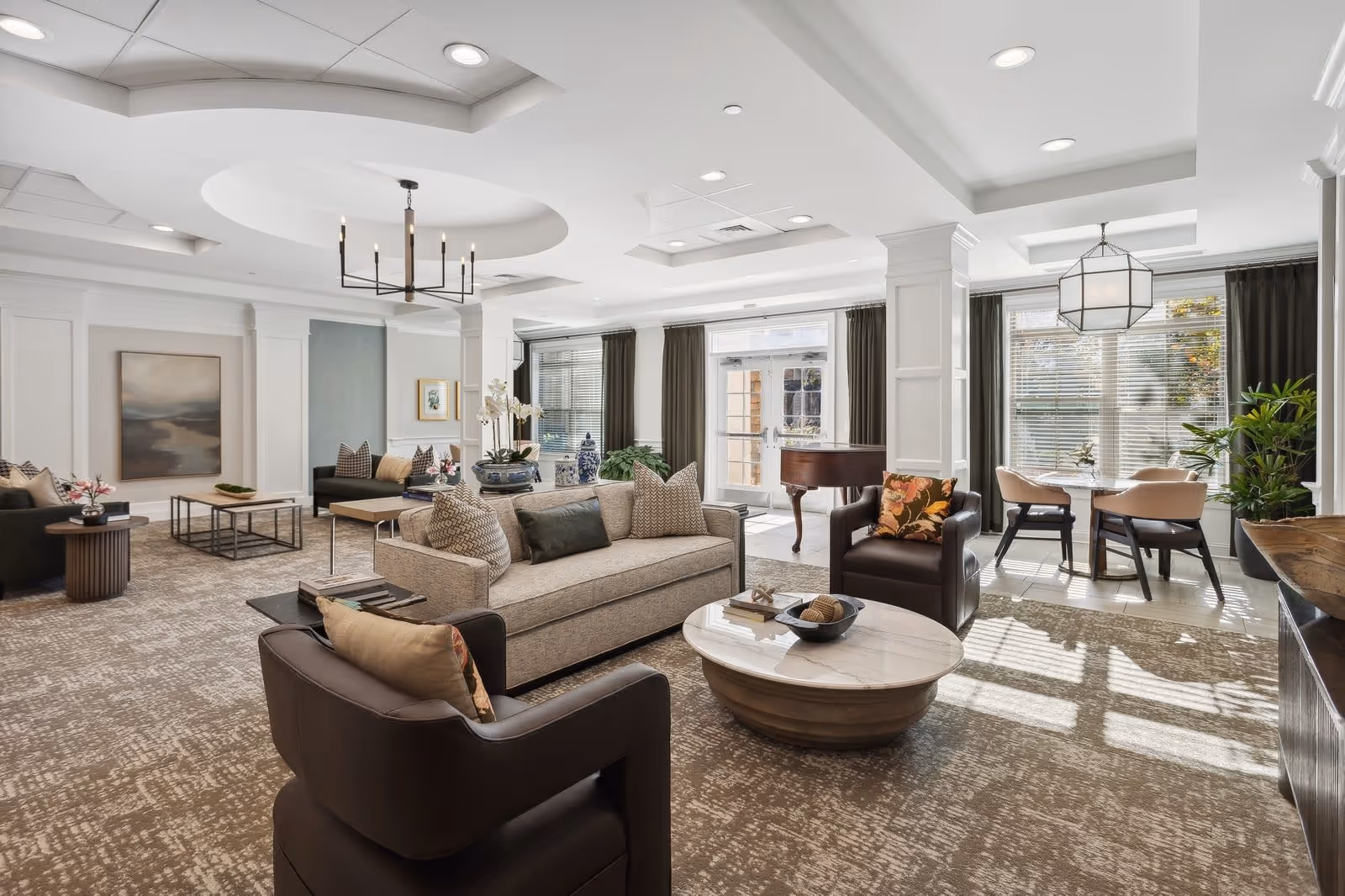 A spacious and well-lit senior living facility common area featuring comfortable seating including sofas and armchairs arranged around a round coffee table. Large windows with dark curtains allow natural light to fill the room. There is a grand piano near the windows and a small dining area with a table and chairs. The room has elegant ceiling details and modern light fixtures.