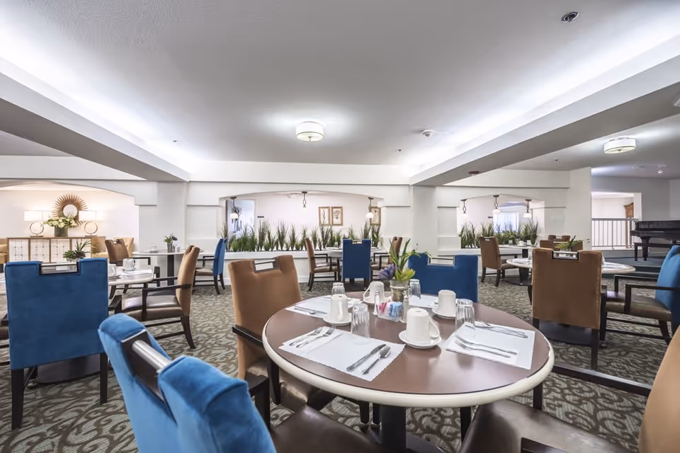 Bright communal dining room with round tables set for meals, upholstered blue and brown chairs, and decorative plants.