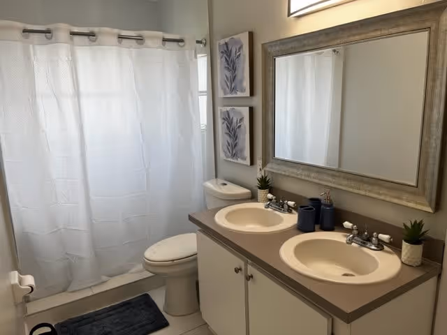 A clean bathroom with a white shower curtain, a toilet, and a double sink vanity with two faucets. Above the sinks is a large rectangular mirror with a wooden frame. Two small potted plants and dark blue soap dispensers are placed on the countertop. Two framed botanical prints hang on the wall above the toilet. A dark blue bath mat is on the floor in front of the shower.