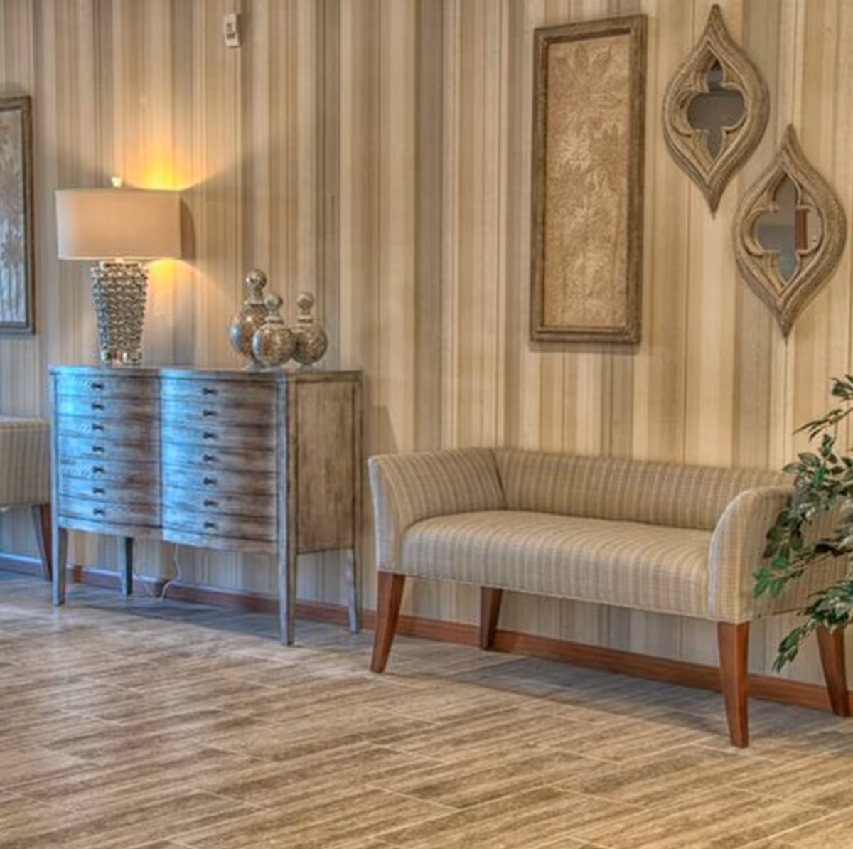 A cozy interior corner featuring a striped upholstered bench with wooden legs, a rustic wooden sideboard with multiple drawers, a decorative table lamp, and ornamental vases. The wall behind has vertical striped wallpaper and is adorned with framed artwork and two uniquely shaped mirrors. A leafy green plant is partially visible on the right side.