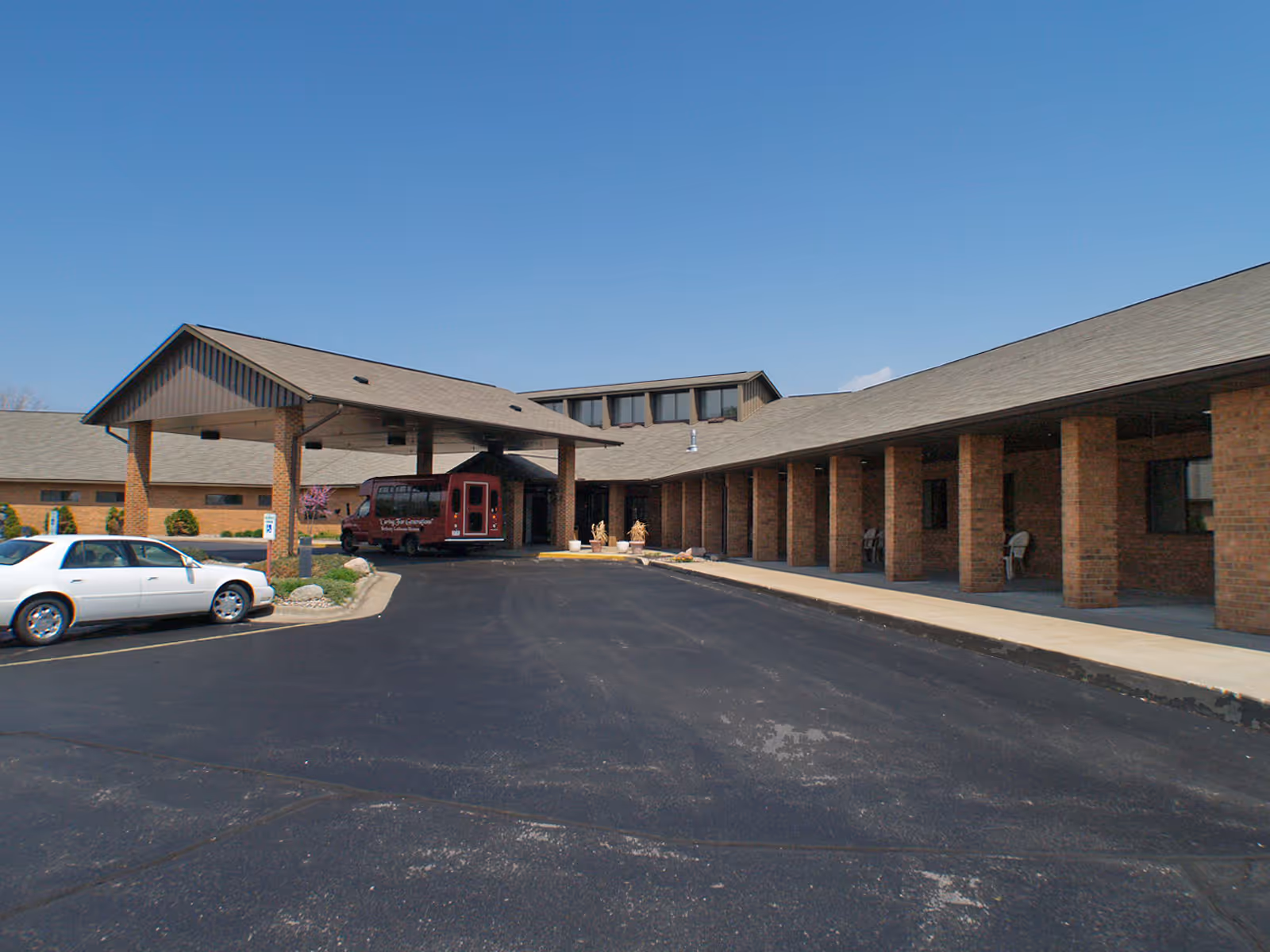 Front exterior of a single-story brick senior care facility with a covered drop-off portico, parking lot, and vehicles.