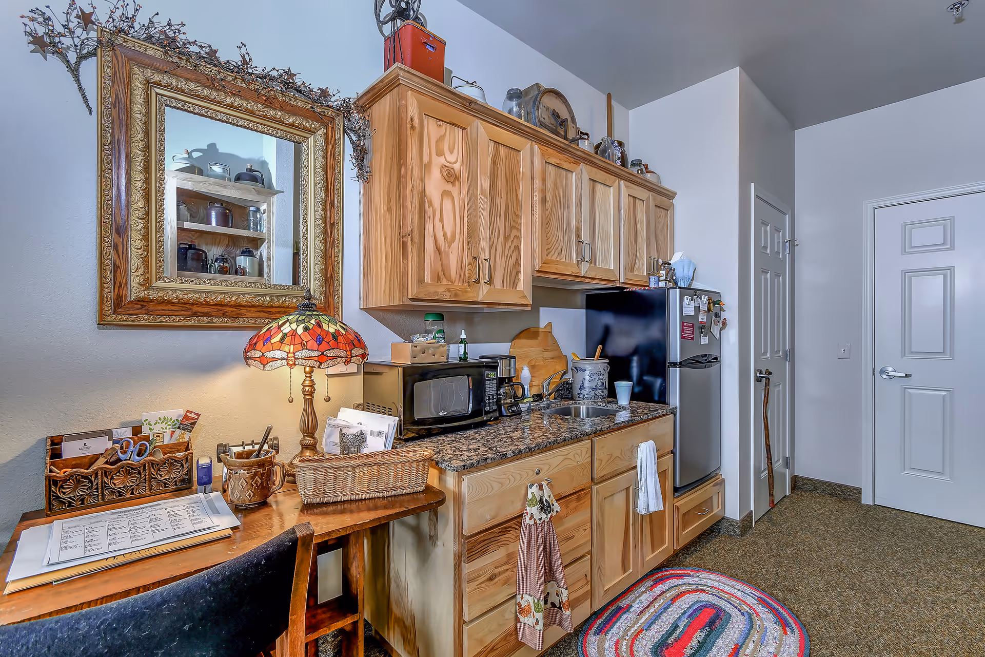 Small kitchenette with wooden cabinets, granite countertop, sink, microwave and mini fridge beside a desk with a decorative lamp and mirror.