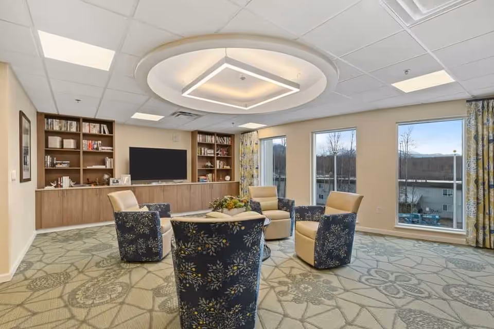 Bright senior living lounge with a circle of upholstered chairs around a central table, built-in bookshelves and a TV beneath large windows.