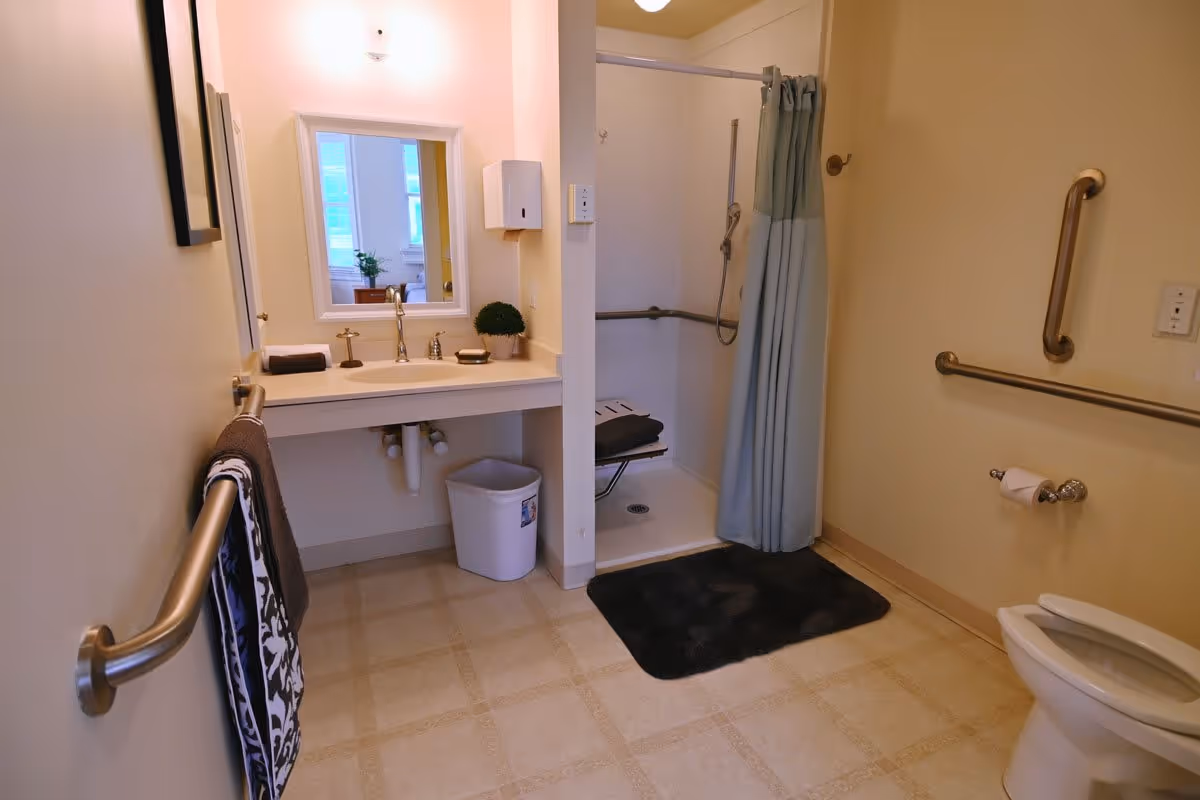 Accessible bathroom with sink and mirror, roll-in shower with seat and curtain, toilet, grab bars, and towel racks.