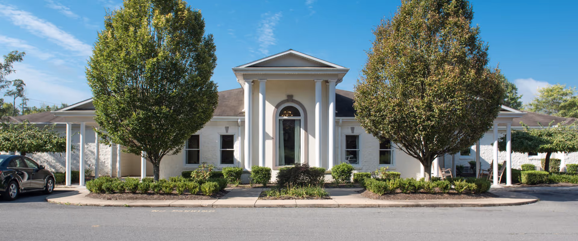 Front exterior of a single-story senior healthcare building with a columned entrance, trees, and a driveway.