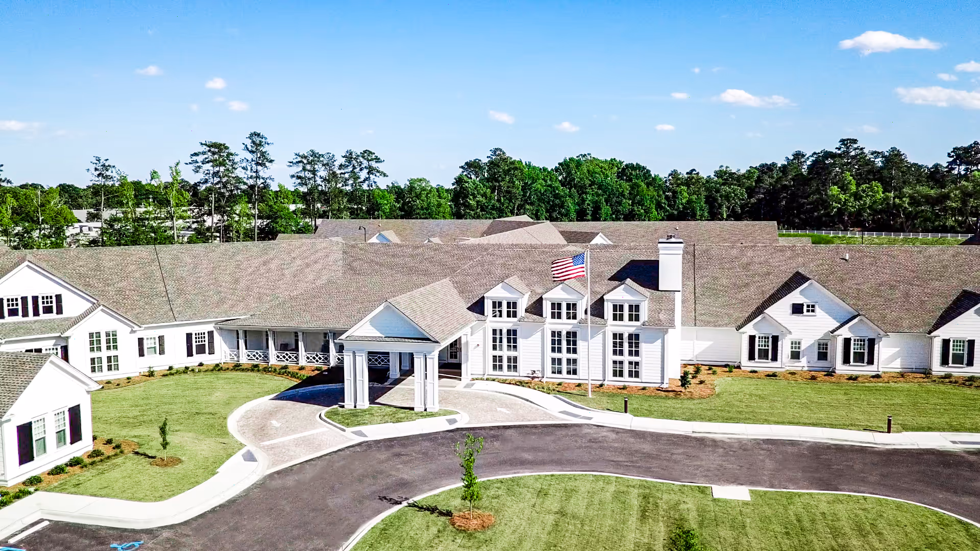 Front exterior of a single-story long-term care facility with a covered entrance, circular drive, and an American flag.