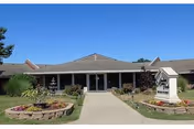 Front entrance of Meadows Manor East: a single-story building with a central walkway, landscaped flowerbeds, and a freestanding sign under a clear blue sky.