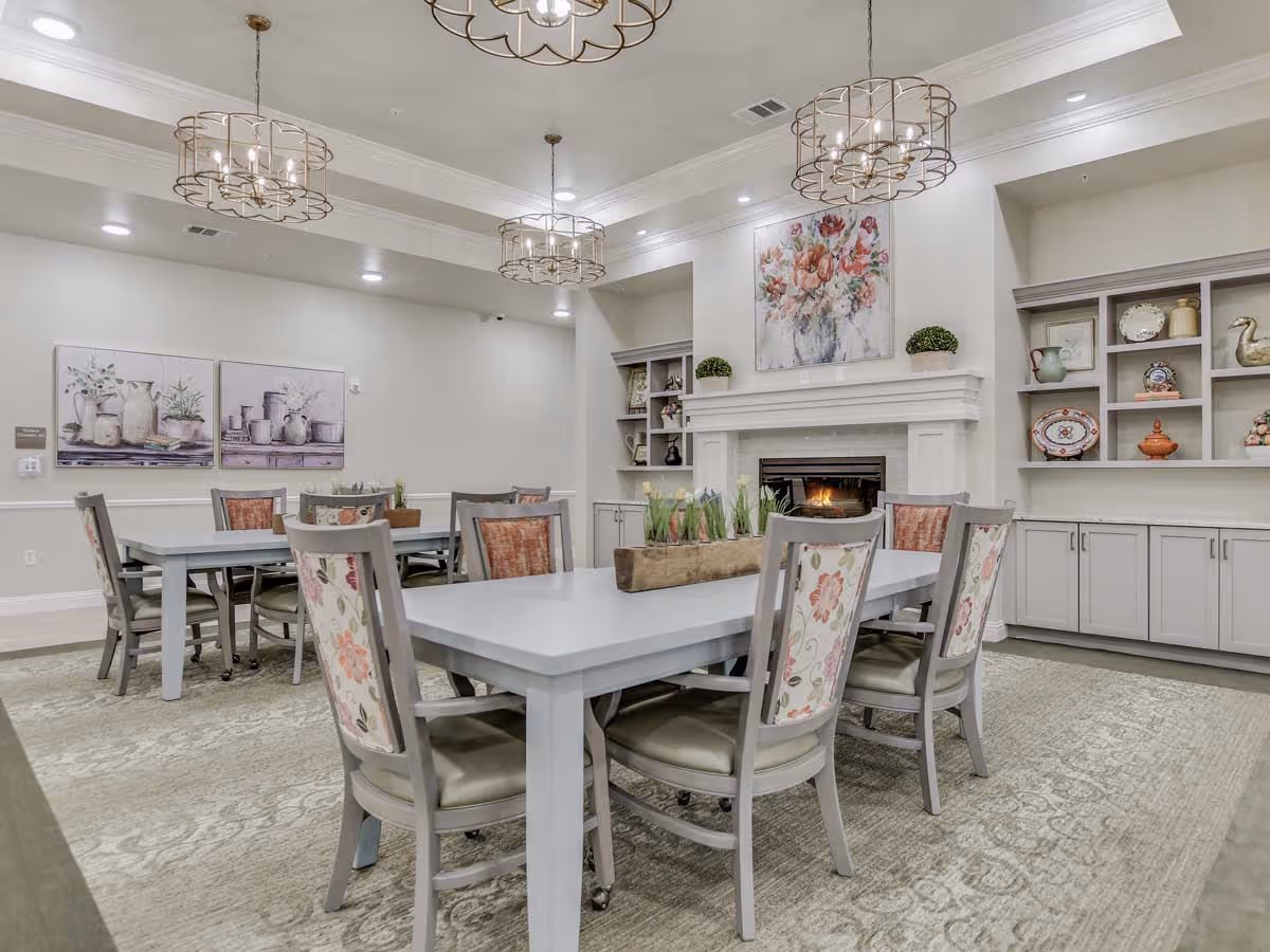 A cozy dining area with two rectangular tables surrounded by chairs with floral upholstery. The room features a fireplace with a floral painting above it, built-in shelves with decorative items, and three modern pendant lights hanging from the ceiling. The walls are painted light gray and the floor is covered with a patterned carpet.