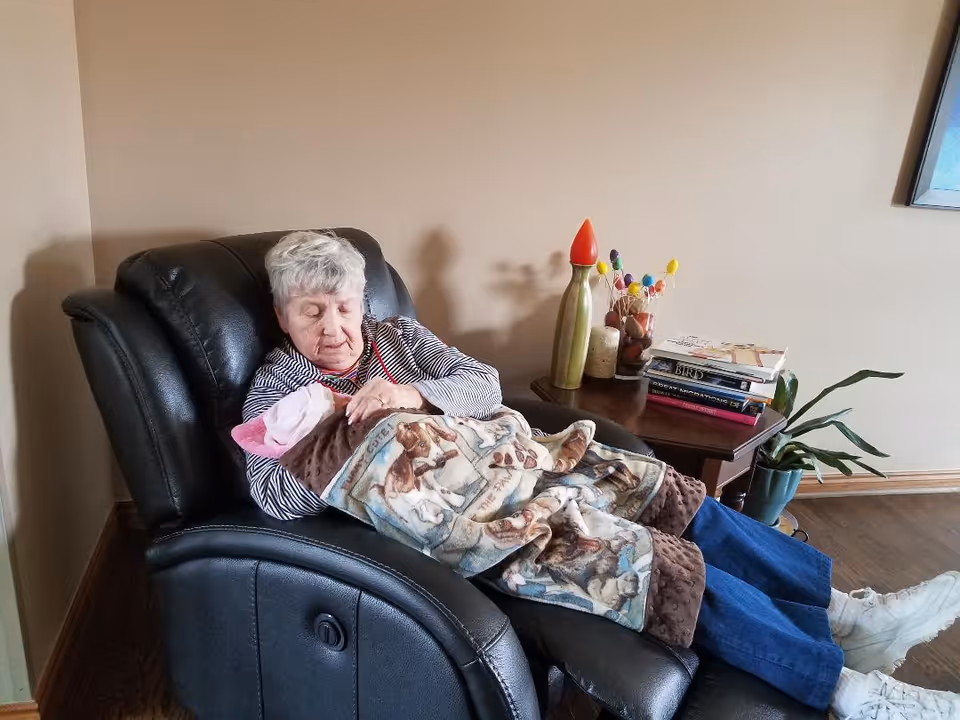 An elderly woman sitting in a black recliner chair holding a baby wrapped in a blanket with dog prints. Next to her is a wooden side table with books, decorative items, and a potted plant on the floor. The room has beige walls and wooden flooring.