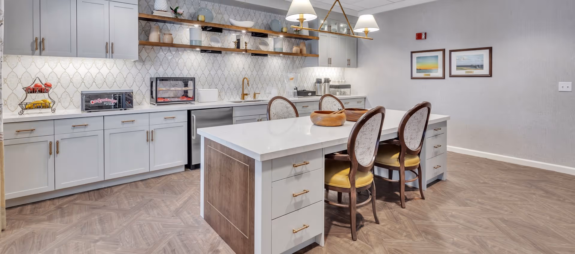 A modern kitchen area with white cabinets, a white countertop island with four chairs, wooden shelves with decorative items, a sink with a gold faucet, and various kitchen appliances. The floor has a wood pattern, and two framed pictures hang on the wall.