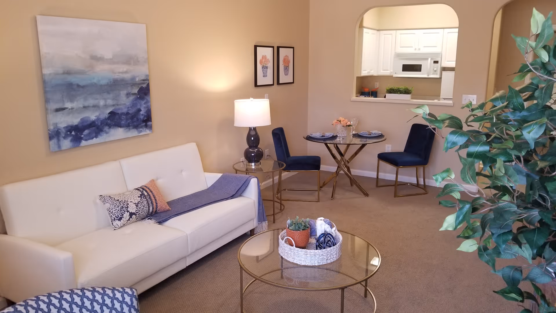 Bright living room with a white sofa, round glass coffee table, small dining set, lamp and wall art, visible through a pass-through into the kitchen.