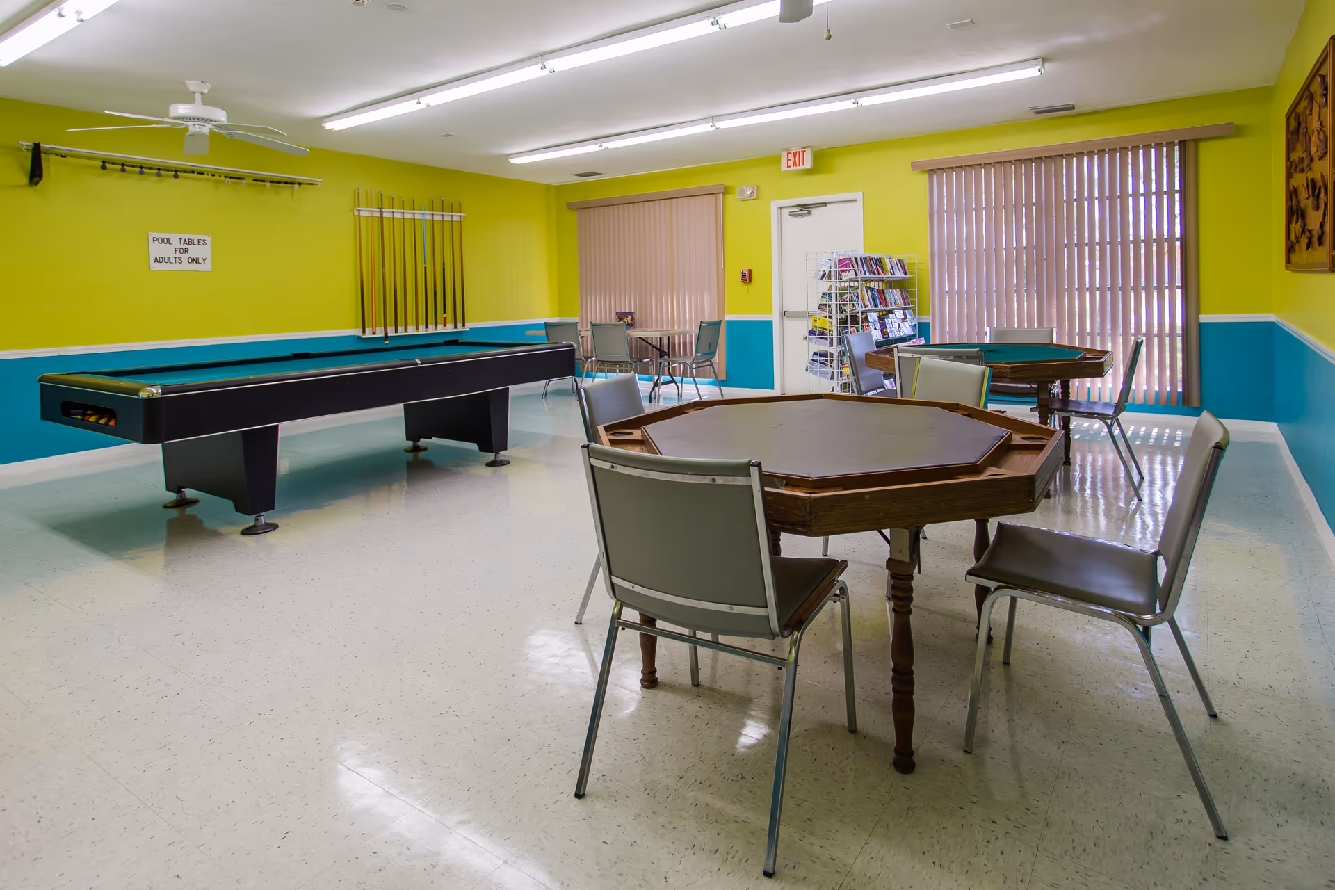 A recreational room with a pool table, several octagonal card tables with chairs, bright yellow and teal walls, a rack of pool cues, and a rack with magazines near a door with an exit sign above it. Vertical blinds cover the windows on the right side.