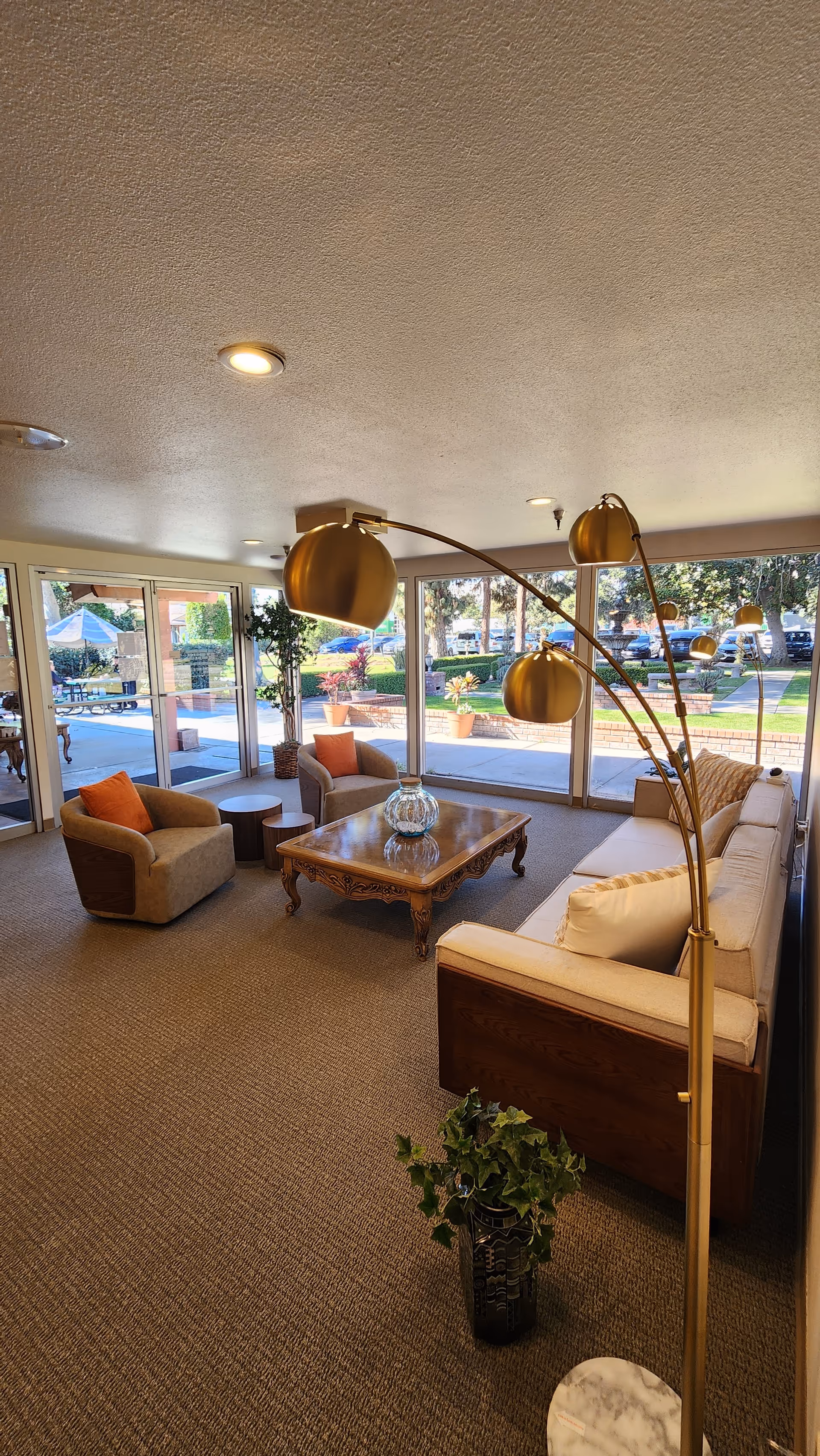 A cozy living room area with a beige sofa, two armchairs with orange cushions, a wooden coffee table with a glass vase, a tall gold floor lamp with multiple lights, and large windows showing an outdoor garden and parking area.