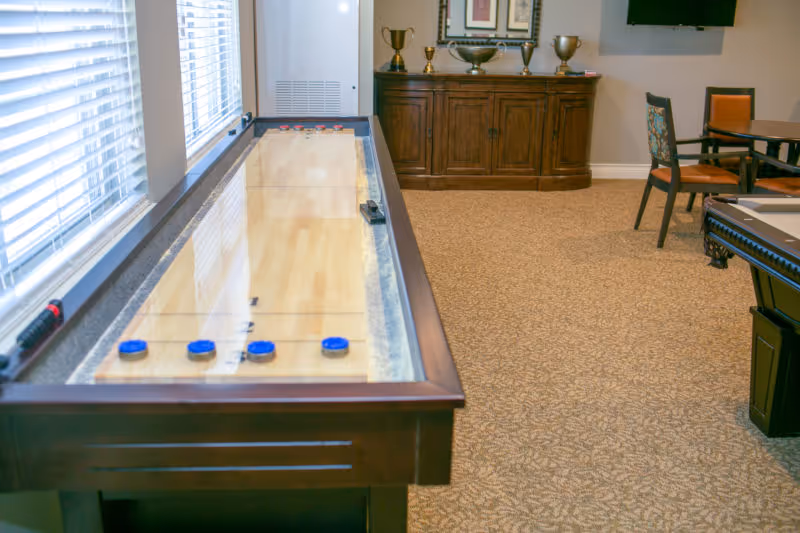 Shuffleboard table in a carpeted senior living common room with chairs, a wooden cabinet, and windows.
