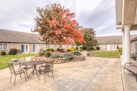 Outdoor courtyard area with a round metal table and four chairs on a concrete patio. There is a raised circular brick planter with a tree that has red and green leaves, surrounded by shrubs. The courtyard is enclosed by a single-story building with multiple windows and doors. The sky is partly cloudy.