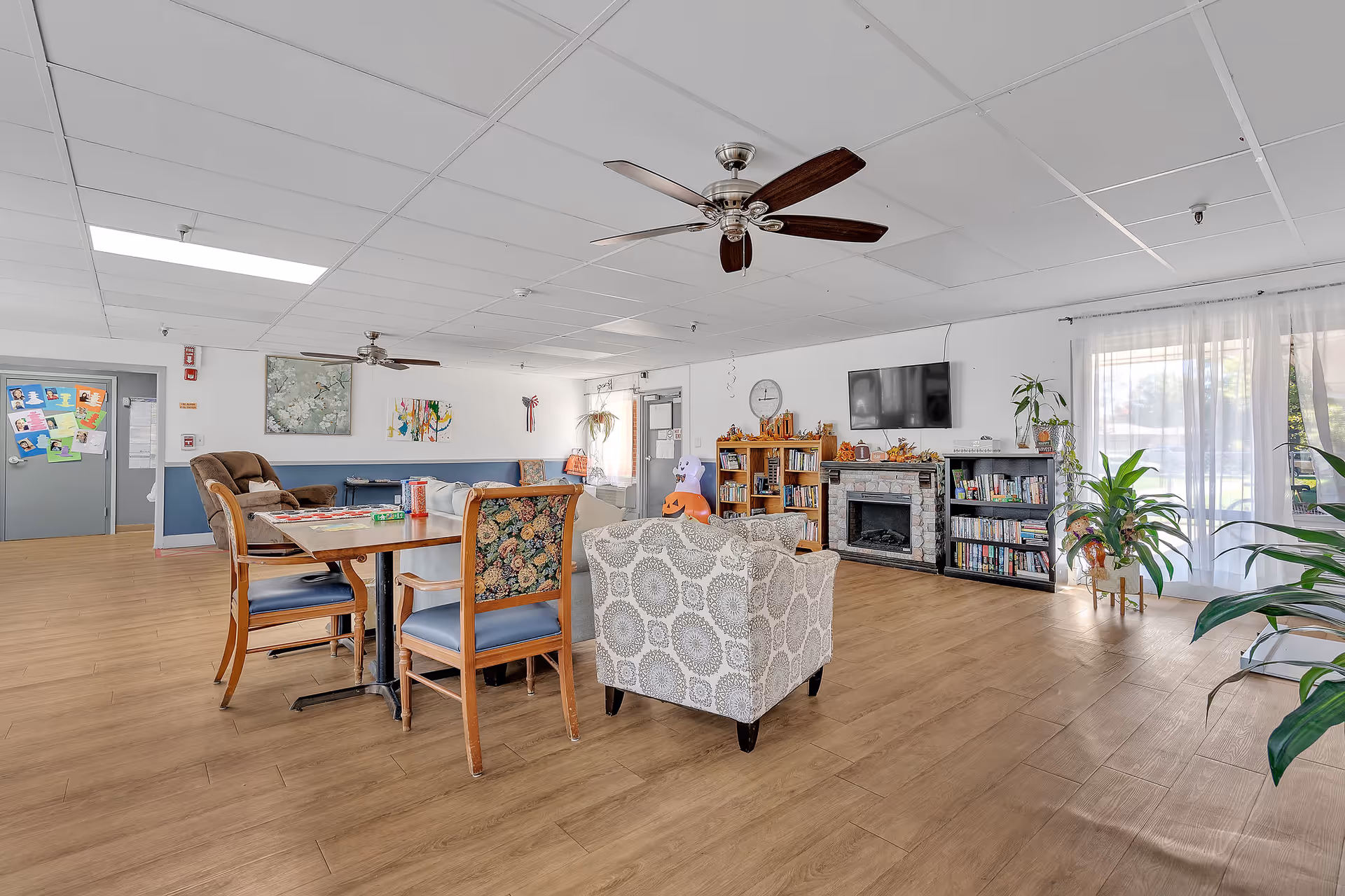 Bright communal living room with seating, a dining table, TV over a fireplace, bookshelves and plants.