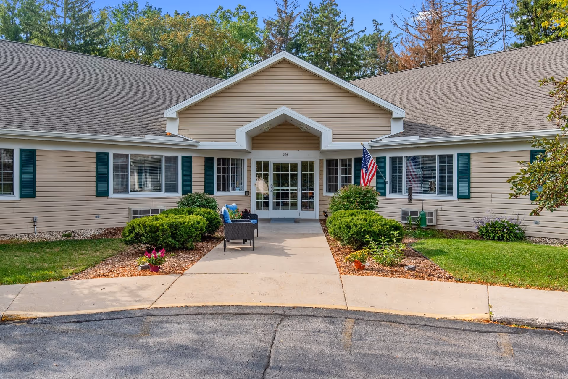 Front exterior view of a single-story beige building with green shutters and a peaked roof entrance. There are bushes and flowers along the walkway leading to double glass doors. An American flag is displayed near the entrance, and two black chairs with blue cushions are placed on the left side of the walkway. Trees are visible in the background under a clear blue sky.