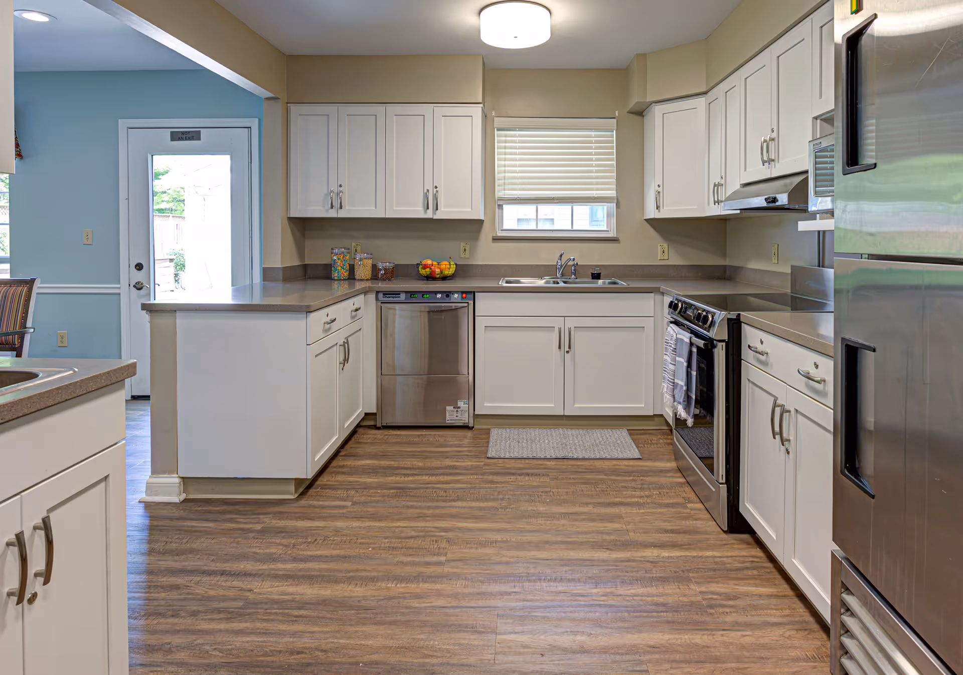 A clean and modern kitchen with white cabinets, stainless steel appliances including a refrigerator, dishwasher, and stove. The kitchen has a window with blinds above the sink, wood-style flooring, and neutral-colored countertops. There is a door leading outside visible in the background to the left.
