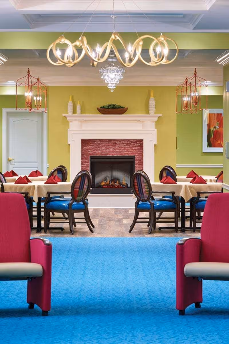 A cozy dining area with tables covered in beige tablecloths and red folded napkins. The chairs have dark wooden frames with blue seats and striped backs. In the background, there is a white fireplace with a red brick interior and decorative vases on the mantel. The walls are painted green, and there are two red hanging light fixtures on either side of the fireplace. The floor has a mix of blue carpet and wood-like tiles. Two red armchairs are partially visible in the foreground.