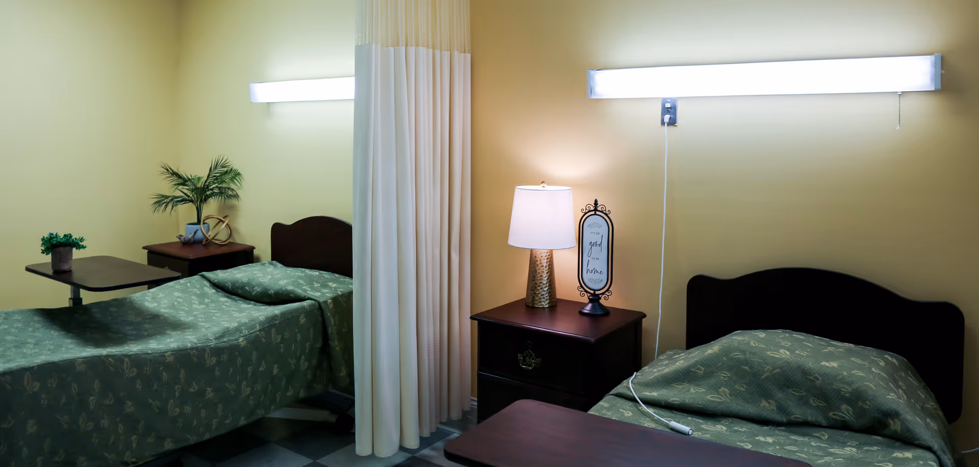 A shared bedroom in a healthcare facility with two single beds separated by a white privacy curtain. Each bed has a green patterned bedspread. There are bedside tables with a lamp, a small plant, and a decorative item. The walls are painted light yellow and there are two long fluorescent lights above each bed.