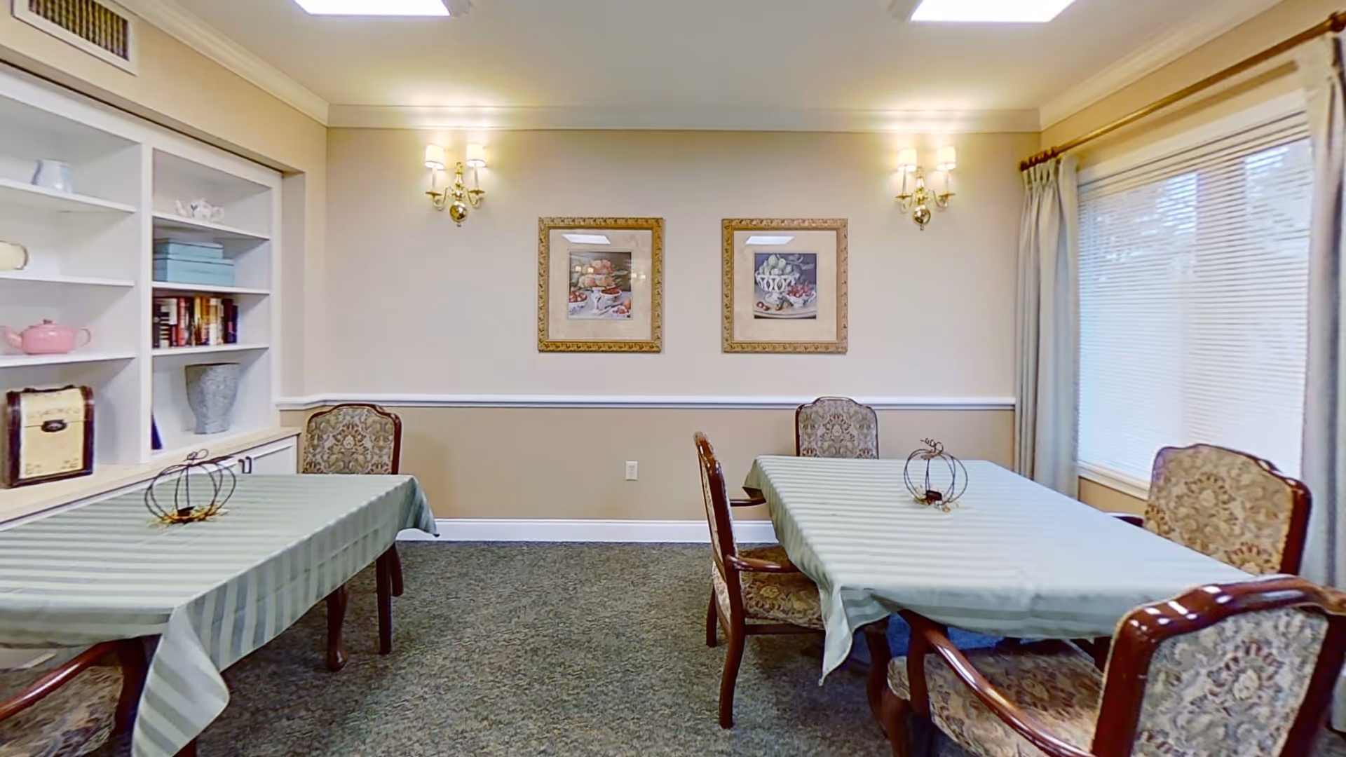 A dining room with two rectangular tables covered with light green striped tablecloths, surrounded by upholstered chairs with floral patterns. The room has beige walls with white trim, two framed pictures on the wall, wall-mounted light fixtures, a large window with blinds and curtains, and built-in shelves with decorative items and books.