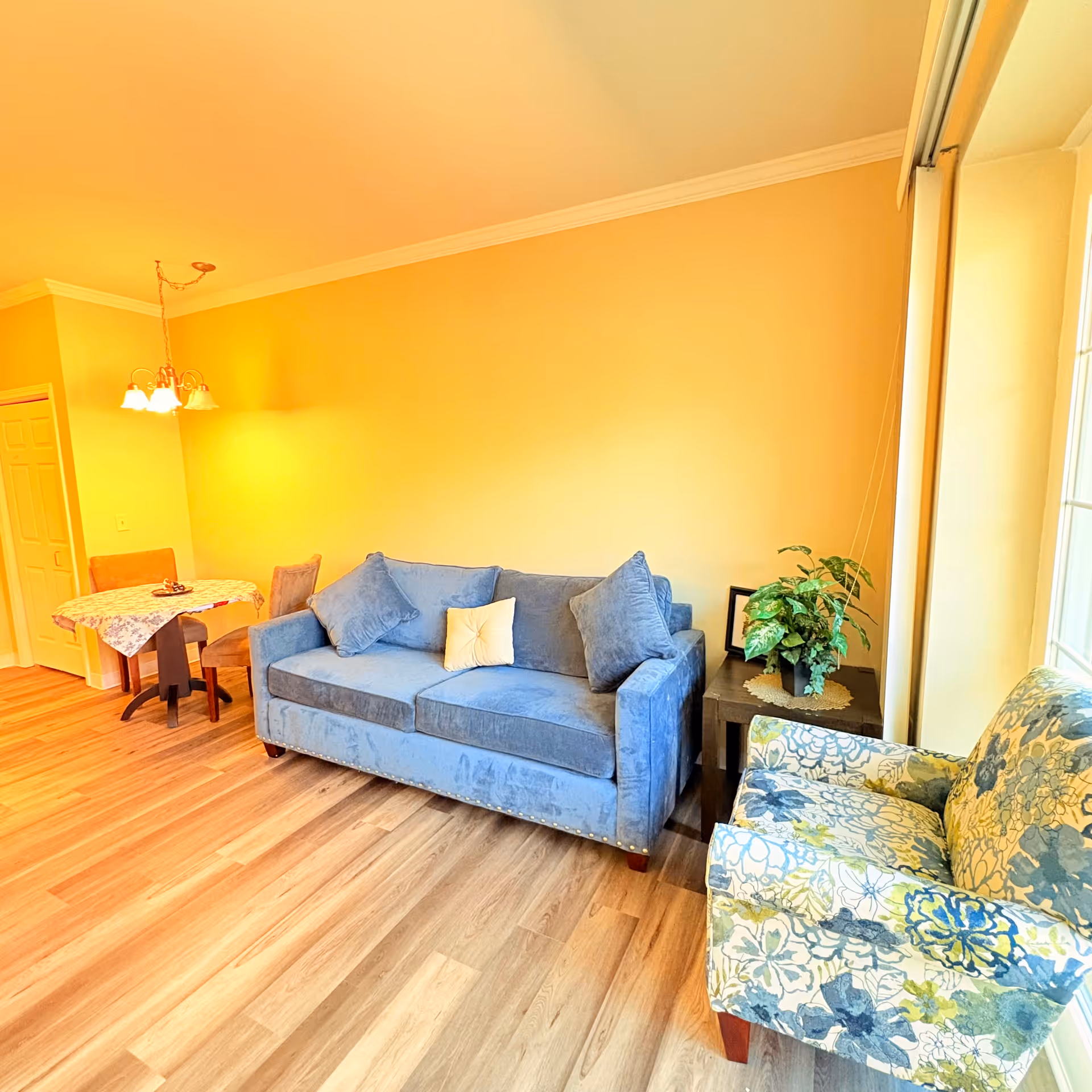 A cozy living area with a blue sofa adorned with three cushions, a floral patterned armchair, and a small side table with a green potted plant and a framed picture. In the background, there is a small dining table with a floral tablecloth and two chairs under a hanging light fixture. The room has light yellow walls and wood flooring, with a large window letting in natural light.