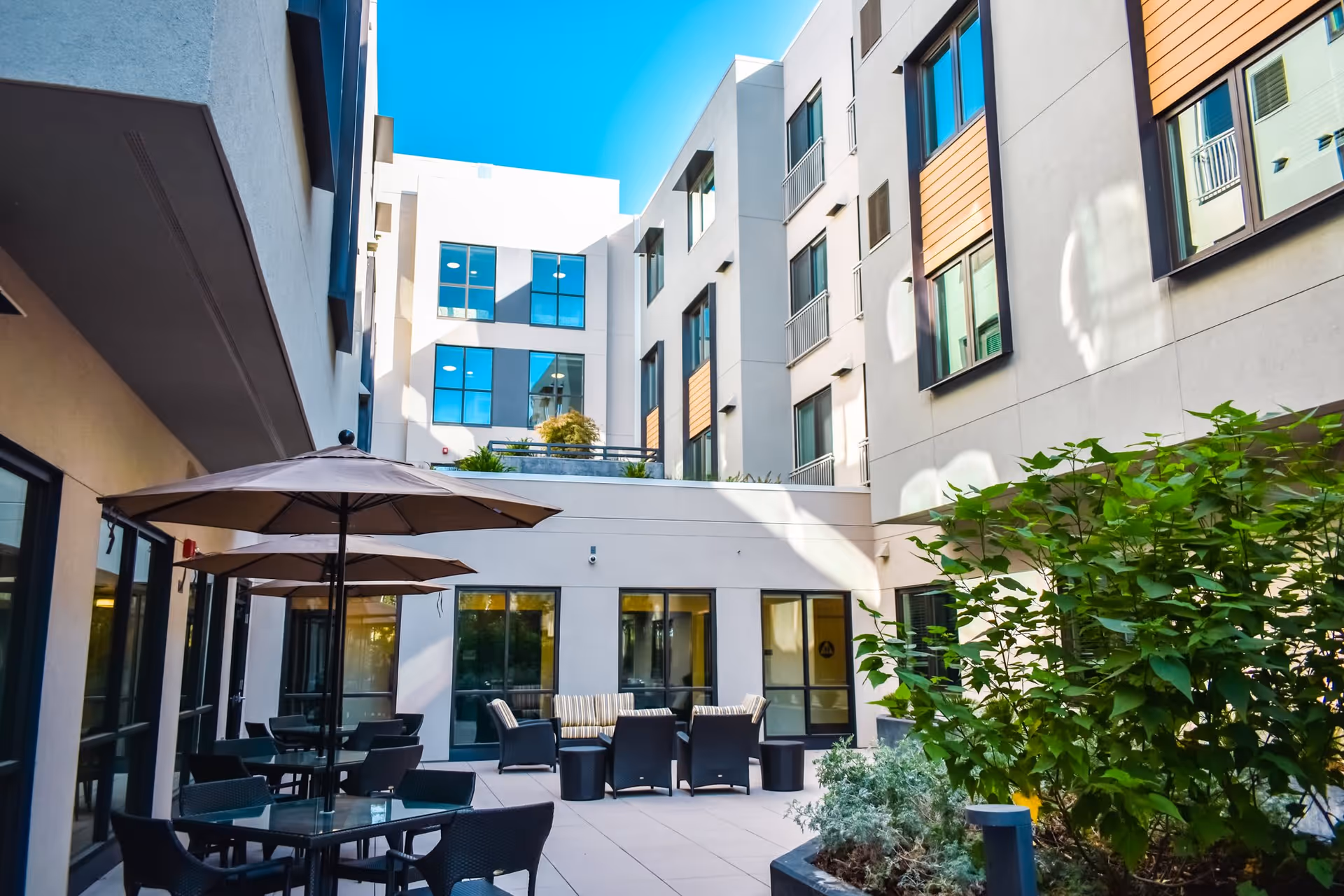 Outdoor courtyard area of a senior living facility with multiple tables and chairs under large umbrellas, surrounded by a multi-story building with large windows and some greenery in planters.