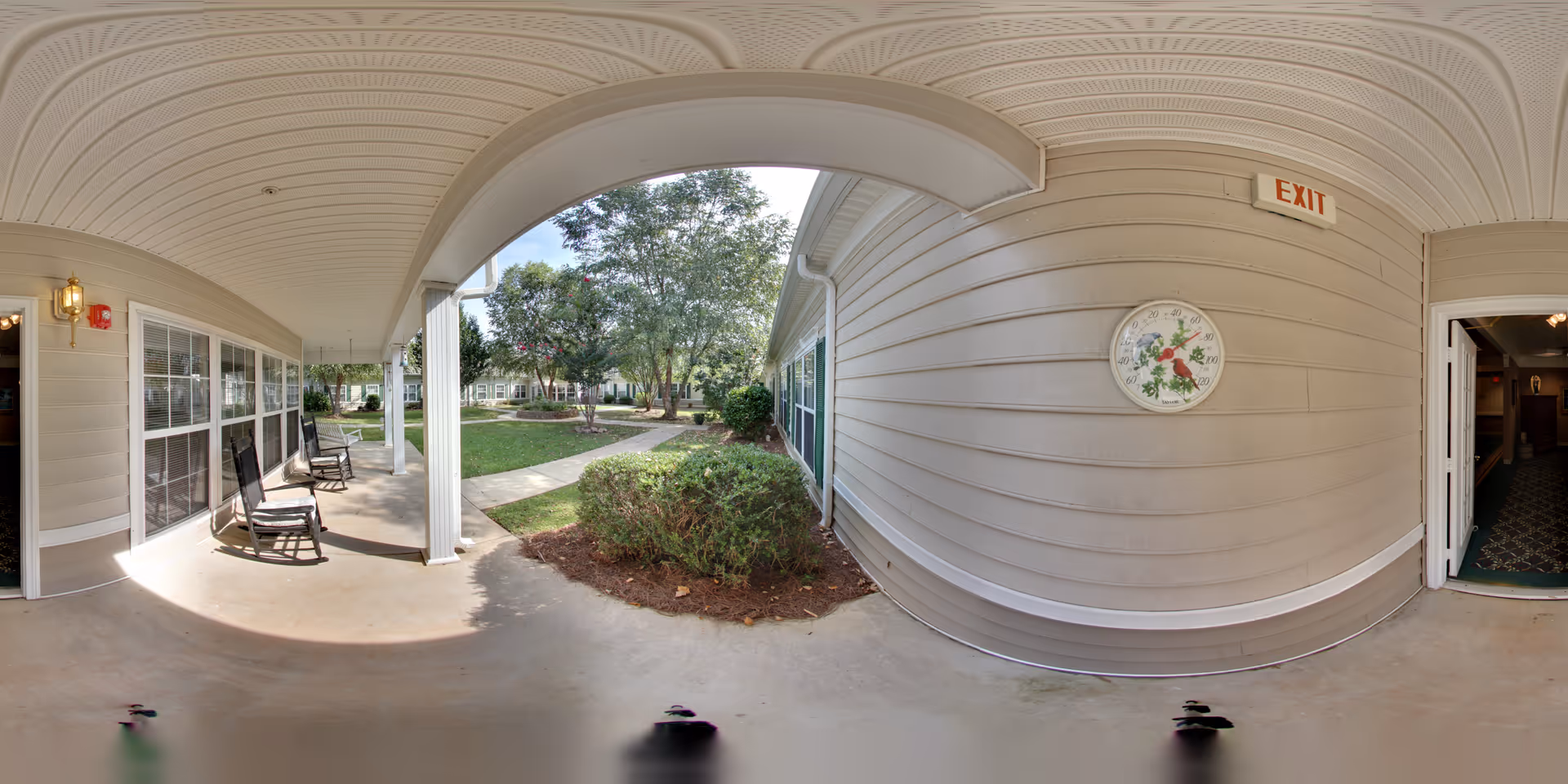 Covered outdoor walkway at Morningside of Anderson with rocking chairs along the wall, a garden area with bushes and trees, a concrete pathway, and an exit sign above a door leading inside.