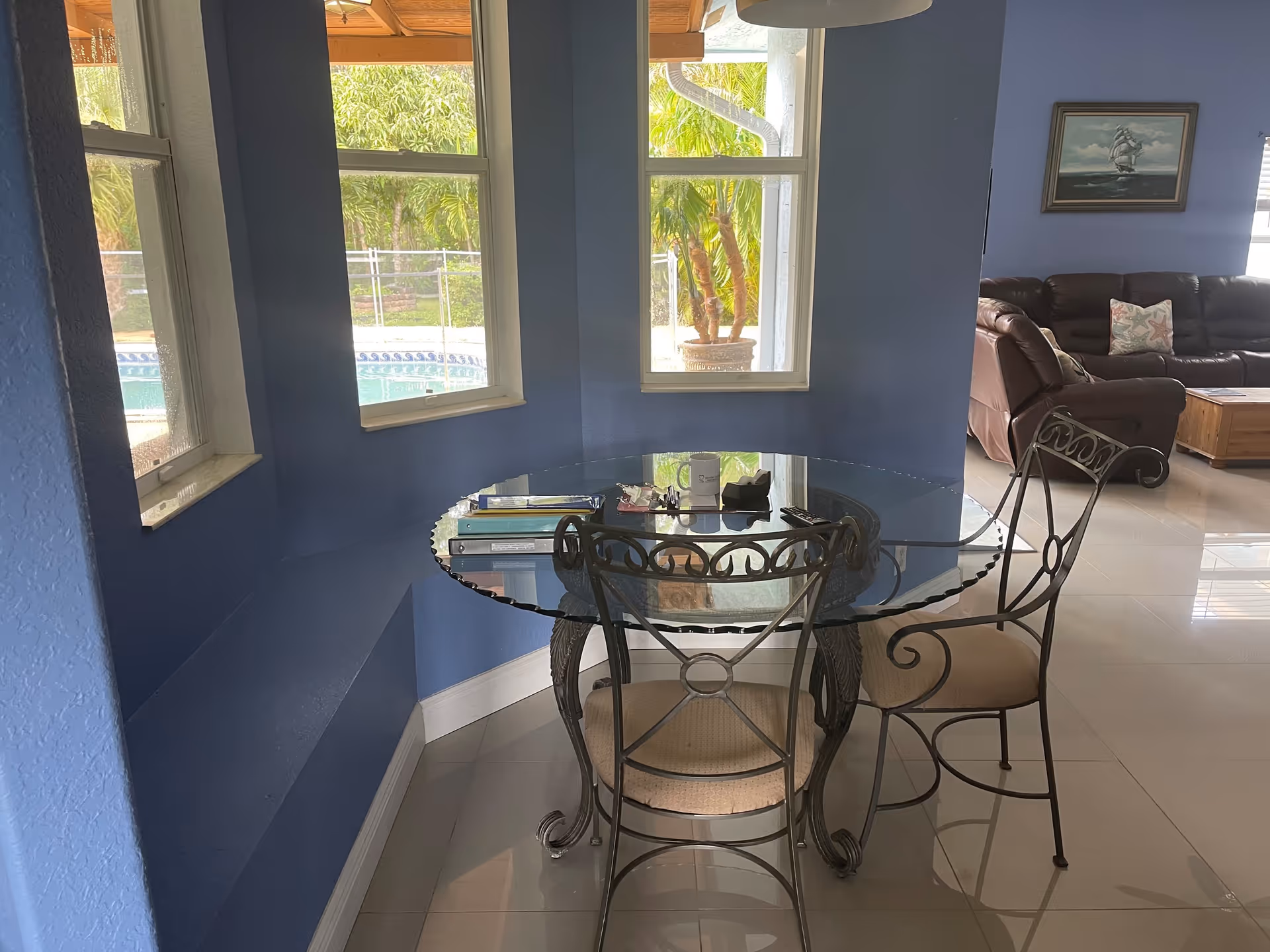 A small dining area with a round glass table and two metal chairs with cushioned seats, positioned near three windows with a view of a swimming pool and palm trees outside. The room has blue walls and a tiled floor, with a living room visible in the background featuring a brown leather sectional sofa, a wooden coffee table, and a painting of a sailing ship on the wall.
