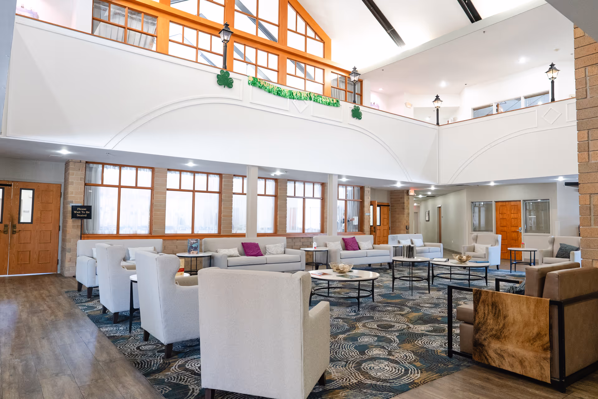 A spacious and well-lit common area in a senior living facility featuring multiple comfortable armchairs and sofas arranged around several round coffee tables on a patterned carpet. The room has large windows with wooden frames, high ceilings with skylights, and decorative wall lamps. There is a balcony area above with green shamrock decorations and a banner.