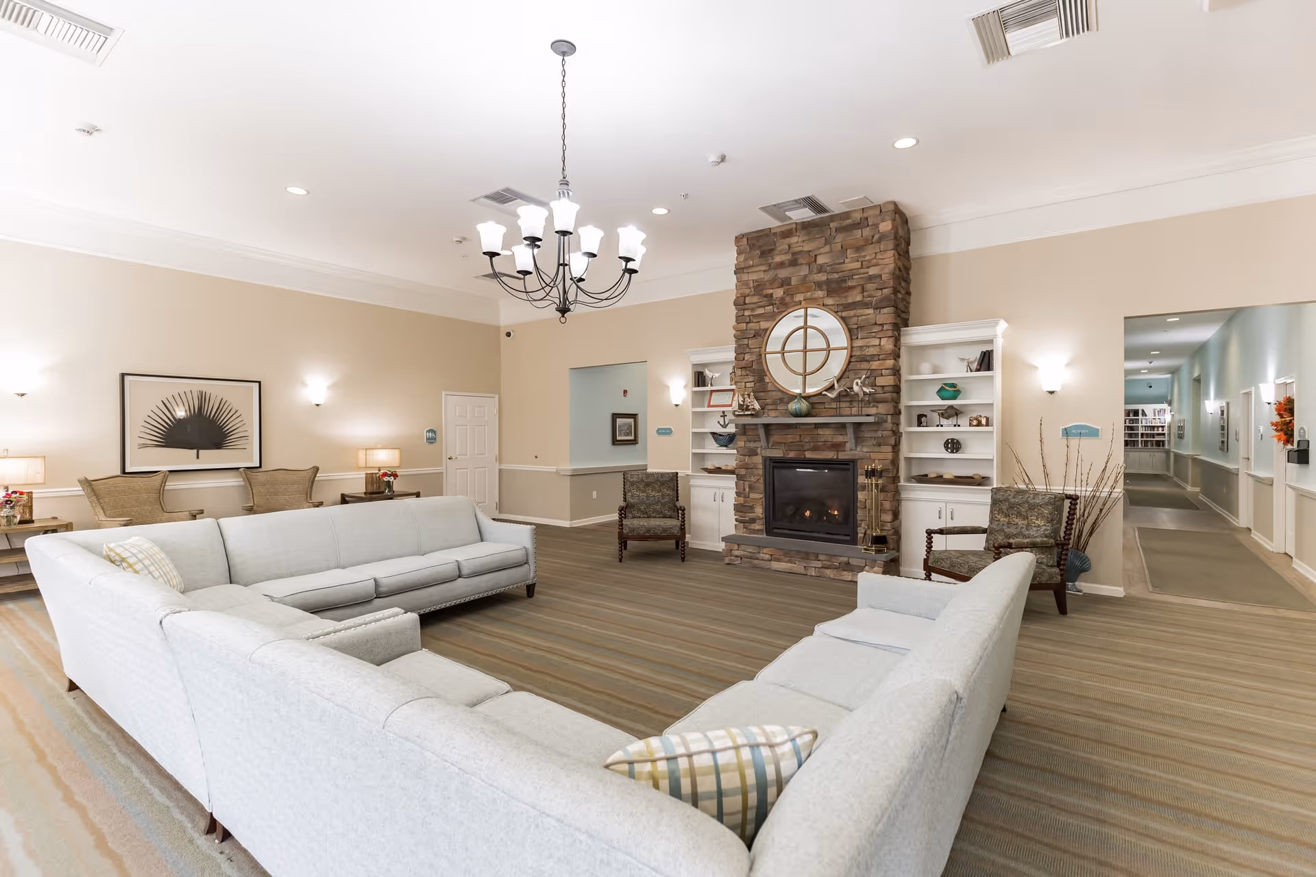 A spacious living room area with a large light gray sectional sofa arranged around a stone fireplace. The fireplace has a round mirror above it and built-in white shelves on either side with decorative items. The room has beige walls, a striped carpet, and a black chandelier hanging from the ceiling. There are two armchairs near the fireplace and a hallway visible to the right.