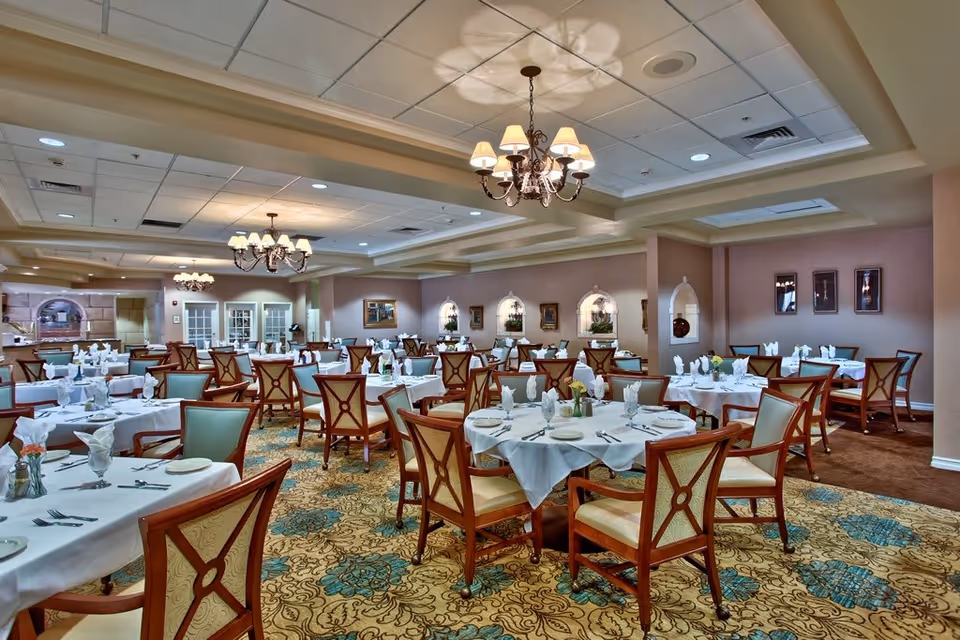 A spacious dining room with multiple round and rectangular tables covered with white tablecloths, set with plates, silverware, and folded napkins. The room features wooden chairs with cushioned seats and backs, patterned carpet with blue and beige floral designs, and elegant chandeliers hanging from a coffered ceiling. The walls are decorated with framed artwork and small alcoves with plants.
