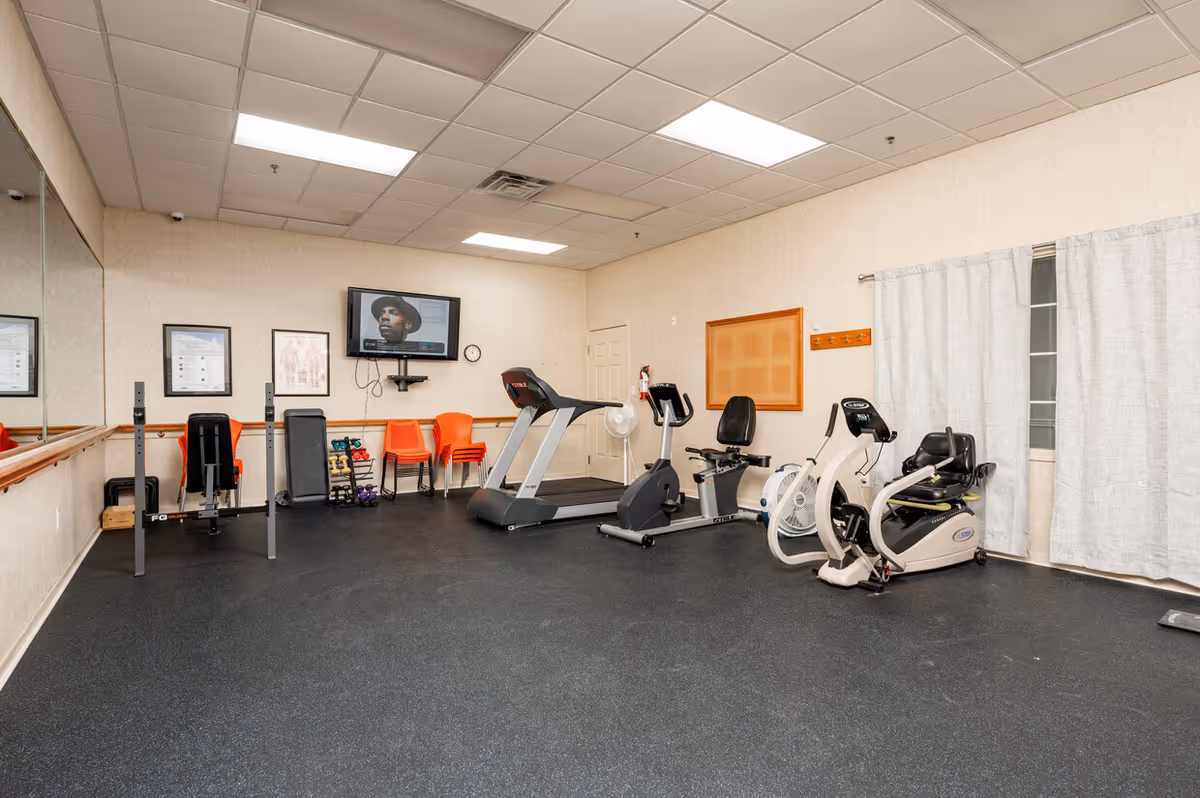 A fitness room with exercise equipment including a treadmill, stationary bike, and weight bench. There are orange chairs stacked against the wall, a wall-mounted TV, a large mirror on one side, and a cork bulletin board on another wall. The room has a black rubber floor and beige walls with a window covered by light-colored curtains.
