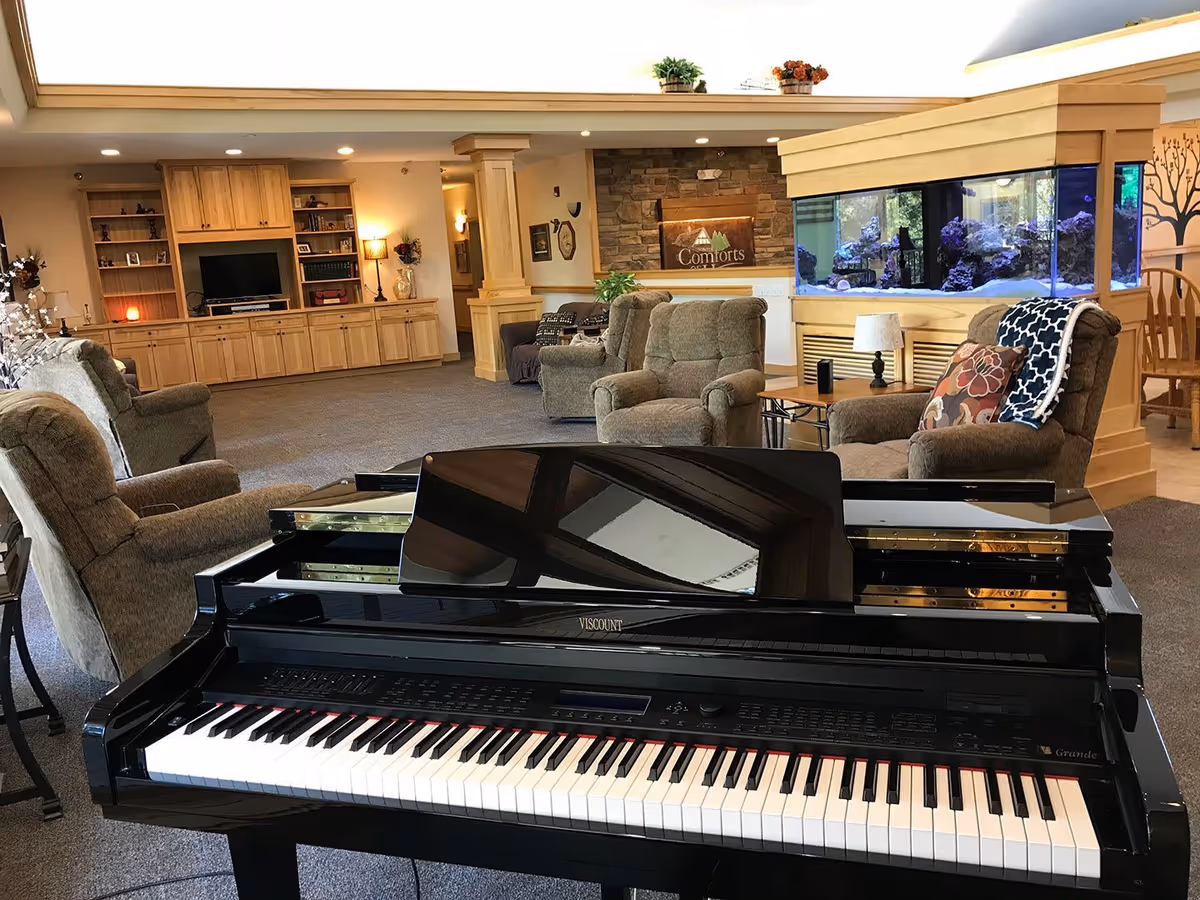 Interior view of a senior living facility common area featuring a black grand piano in the foreground, several comfortable armchairs with cushions and throws, a large built-in wooden entertainment center with a TV, a stone wall with a 'Comforts' sign, and a large aquarium filled with rocks and water. The room is well-lit with ceiling lights and has a cozy, inviting atmosphere.