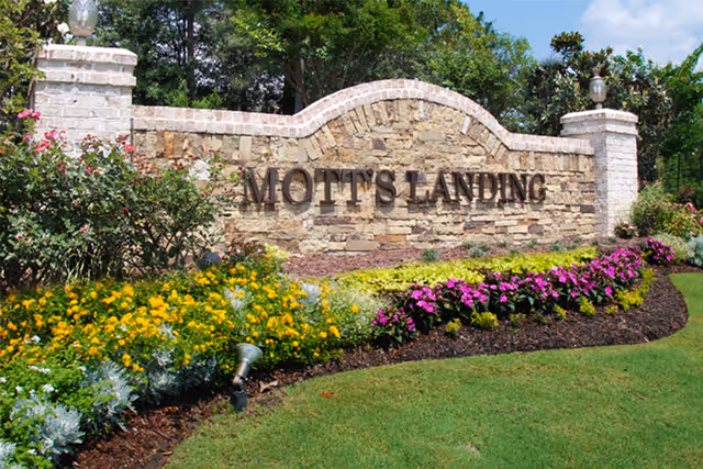 Stone entrance sign for Mott's Landing surrounded by colorful flowers and green grass under a blue sky with some trees in the background.
