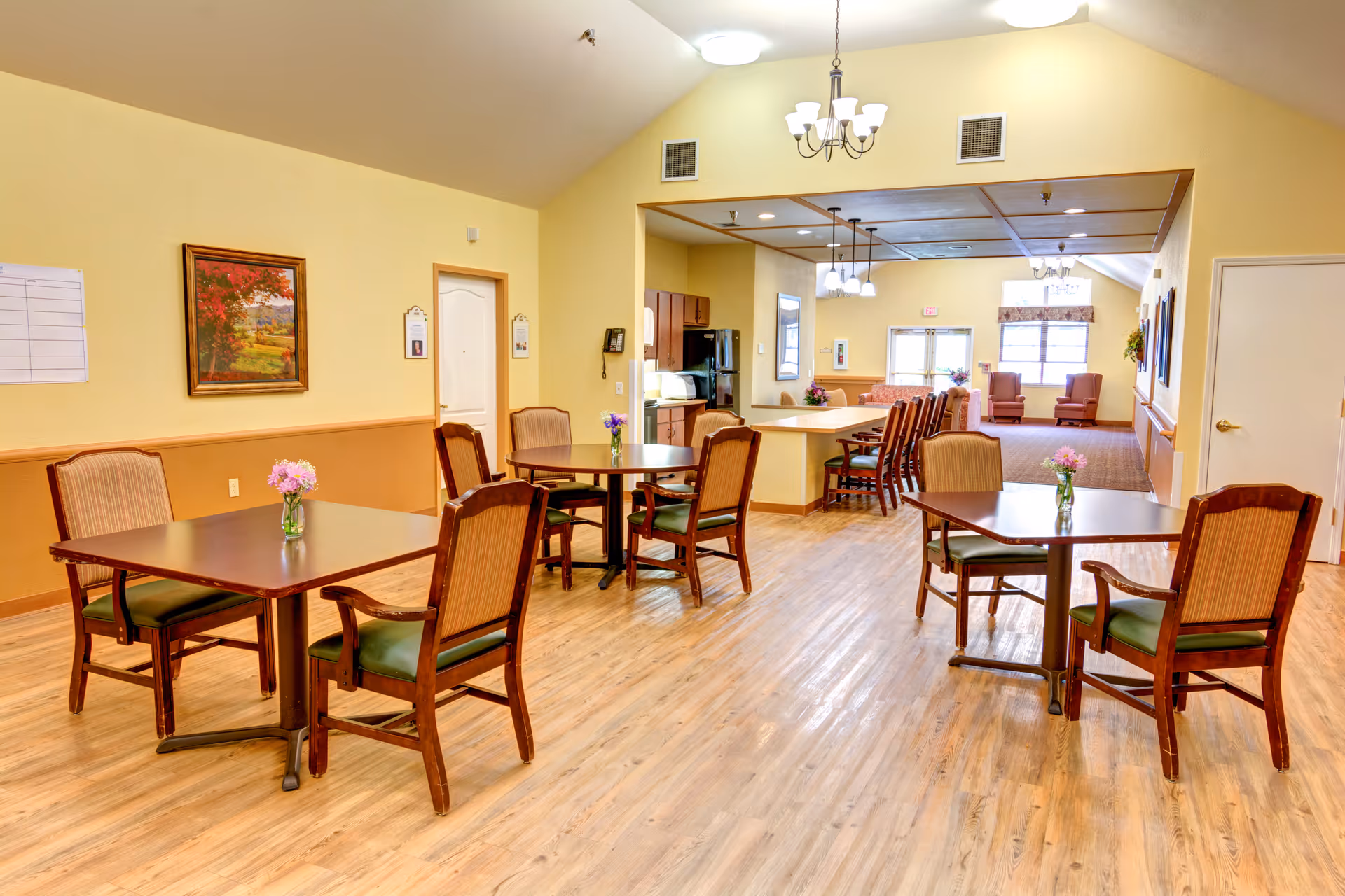 A bright and spacious dining area in a senior living facility with wooden tables and chairs, each table decorated with a small vase of flowers. The room has light yellow walls, wood flooring, and ceiling lights including chandeliers. In the background, there is a kitchen area with a counter and bar stools, and a lounge area with armchairs near windows.