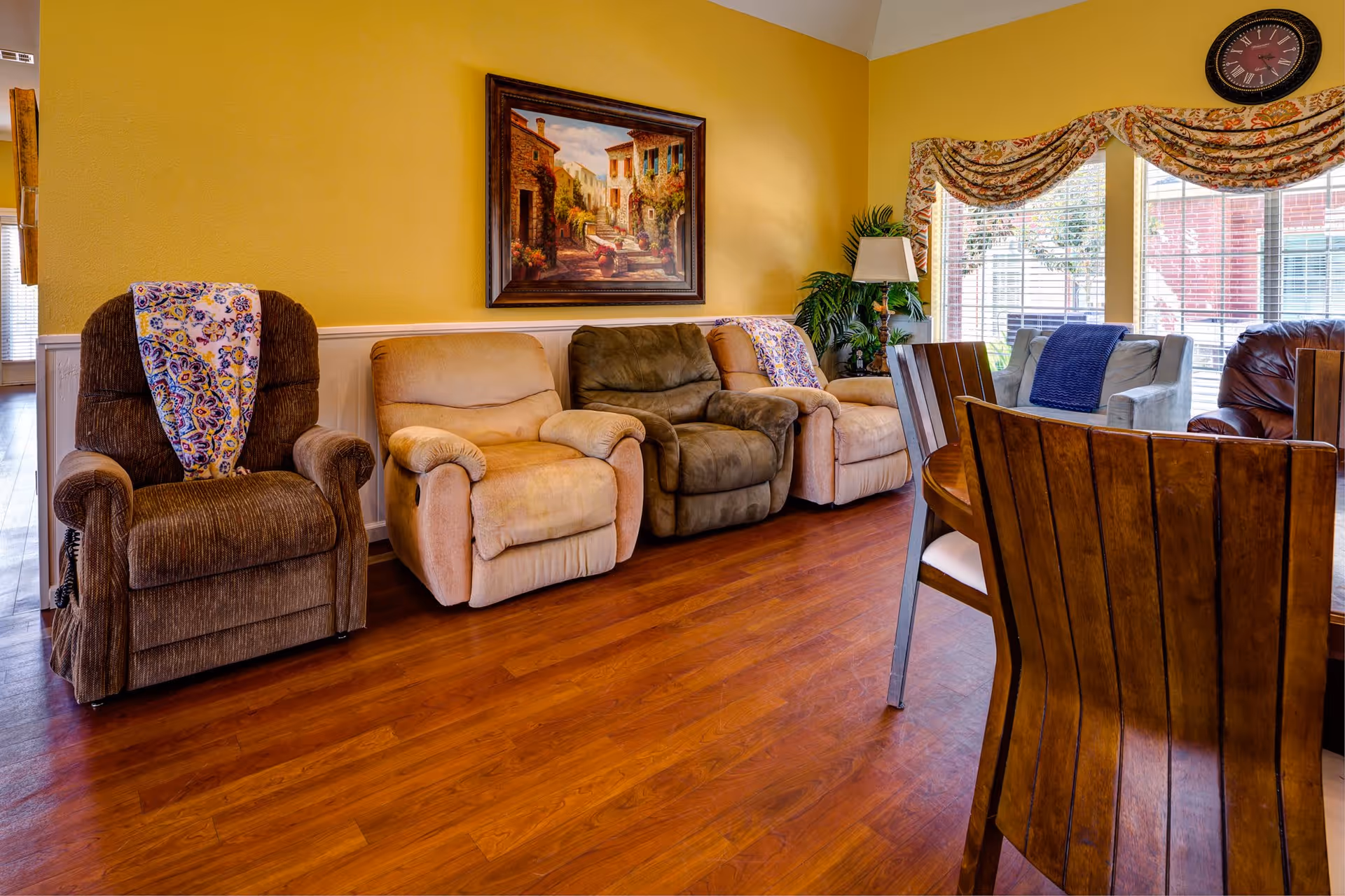 A cozy living room with five recliner chairs in various colors lined up against a yellow wall. Above the chairs is a framed painting of a rustic village scene. To the right, there is a window with floral valances and blinds, letting in natural light. A round wooden table with wooden chairs is partially visible in the foreground, and a potted plant with a lamp is placed near the window.