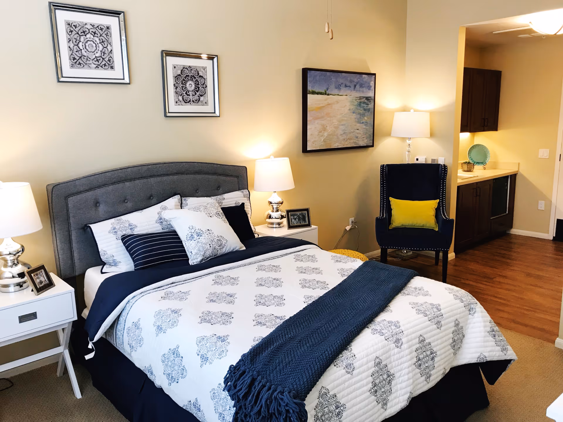 A cozy bedroom with a neatly made bed featuring white and blue patterned bedding and multiple pillows. There are two white nightstands on either side of the bed, each with a silver lamp and framed photos. On the wall above the bed are two framed black and white decorative prints. To the right, there is a navy blue armchair with a yellow cushion, a floor lamp, and a painting of a beach scene on the wall. The room has wooden flooring and an open doorway leading to a kitchenette area with dark cabinets and a countertop.