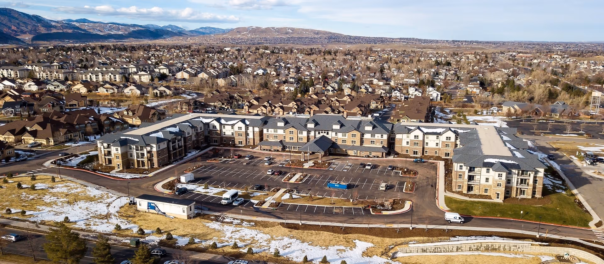 Aerial view of Sky Pointe Retirement Resort, a large multi-story residential building with a central parking lot, surrounded by a suburban neighborhood with many houses and distant mountains under a partly cloudy sky.