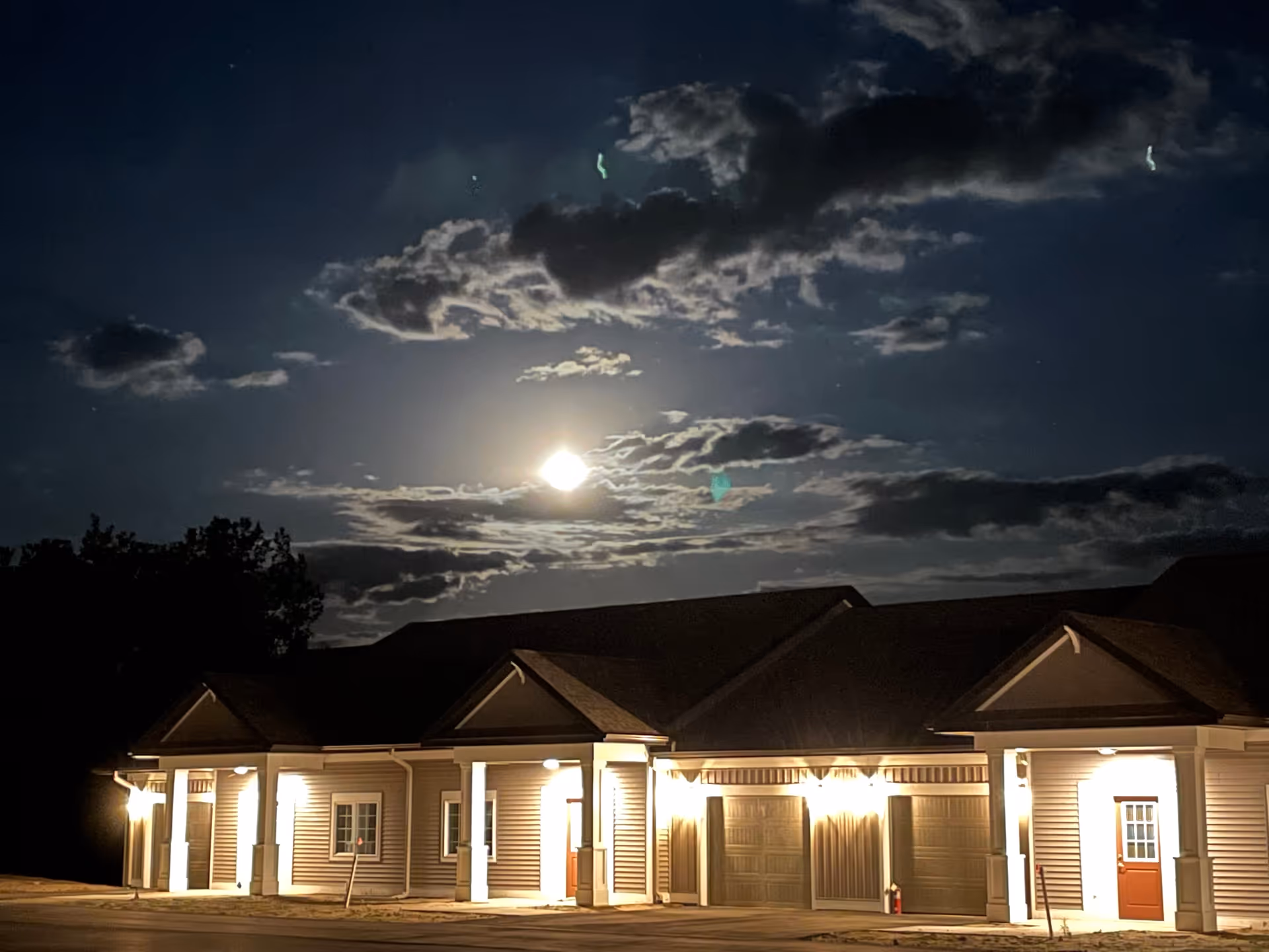 Nighttime view of a single-story building with multiple garage doors and entrances, illuminated by exterior lights under a cloudy sky with a bright full moon.